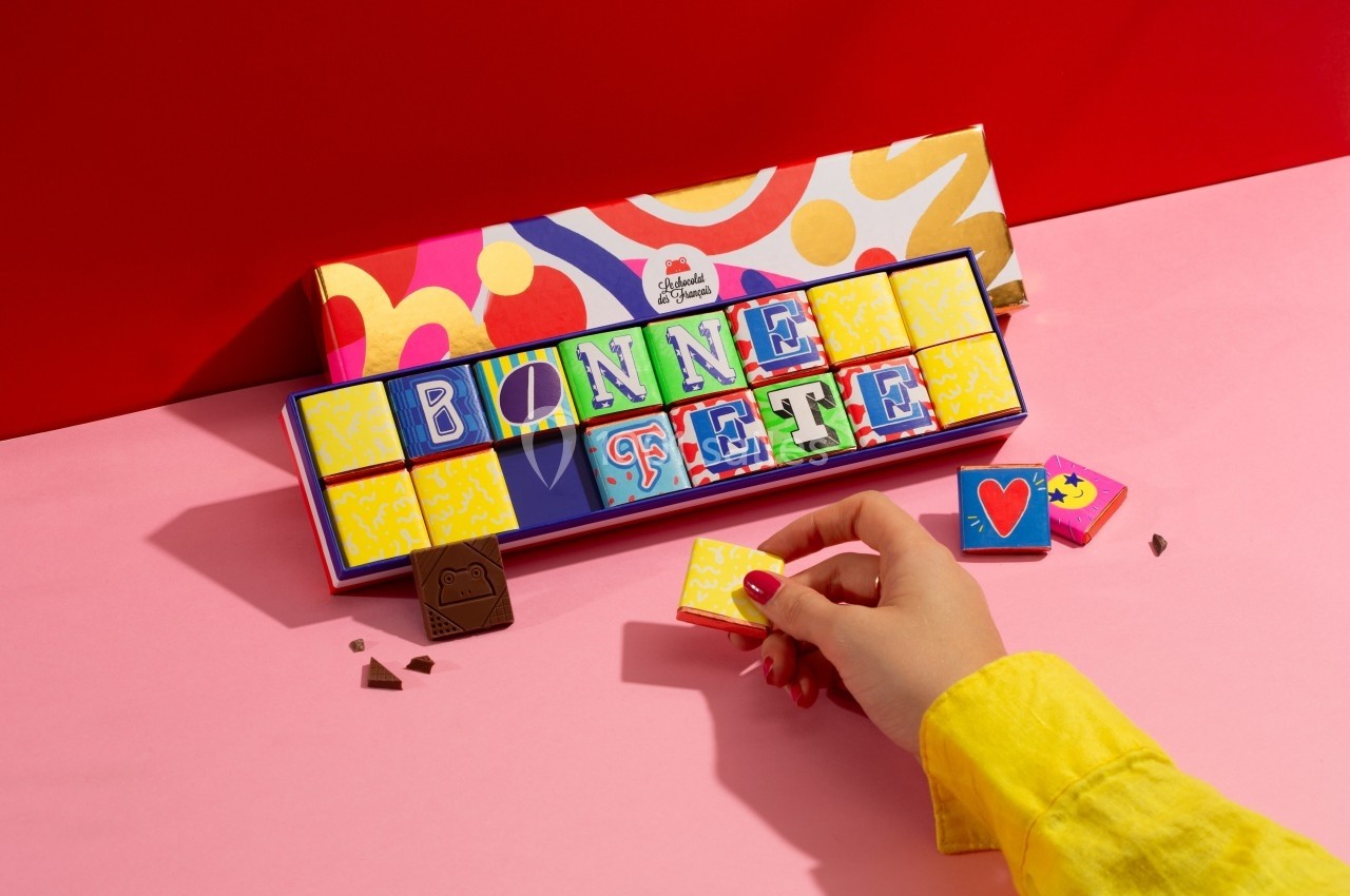 Chocolats colorés formant les mots ’Bonne Fête’ dans une boîte, une main prenant un carré décoré.