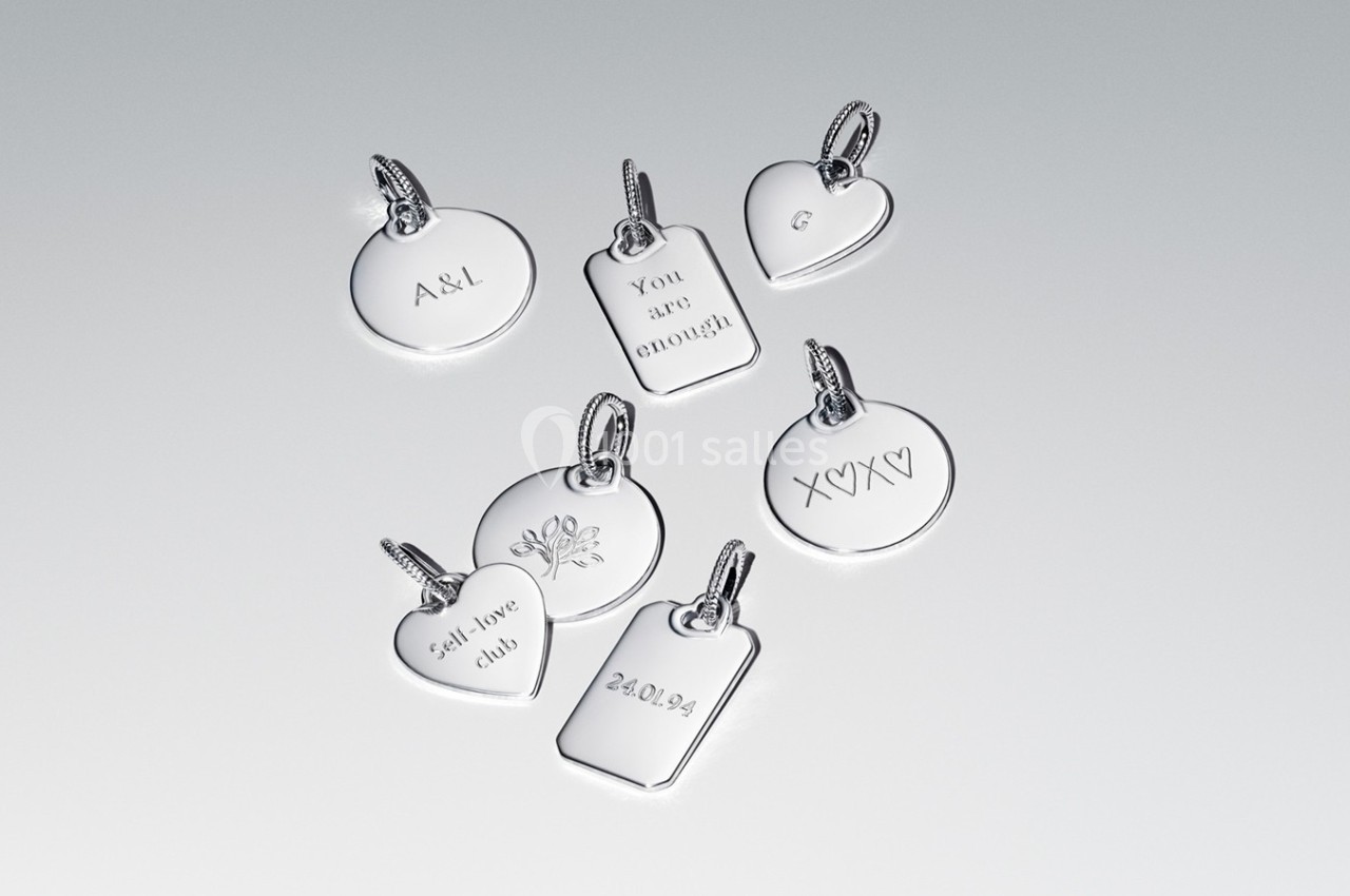 Pendentifs en argent gravés de motifs, initiales, dates et messages, disposés sur une surface claire.