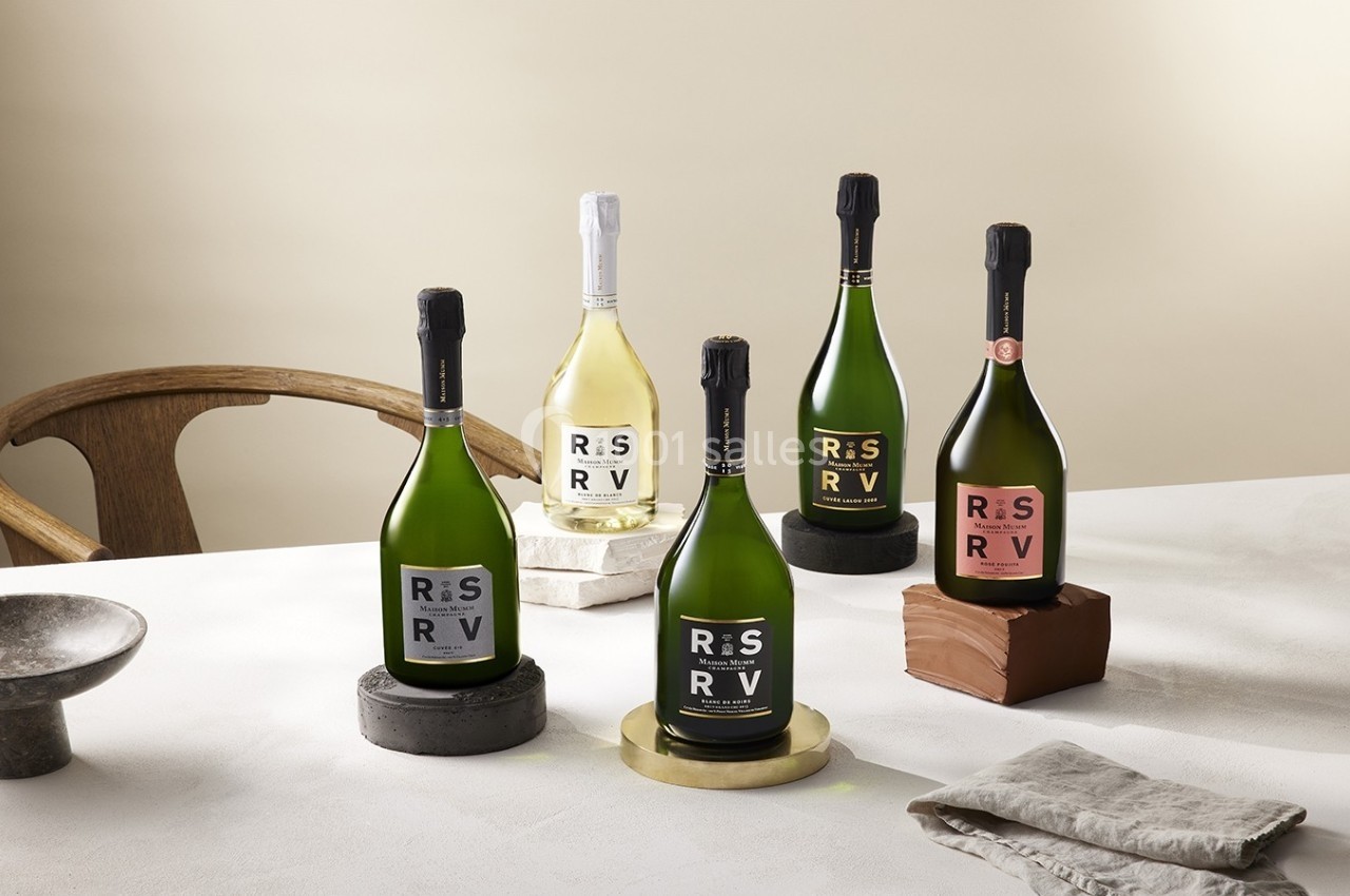 Cinq bouteilles de vin mousseux disposées sur des supports variés sur une table, avec une chaise en arrière-plan.