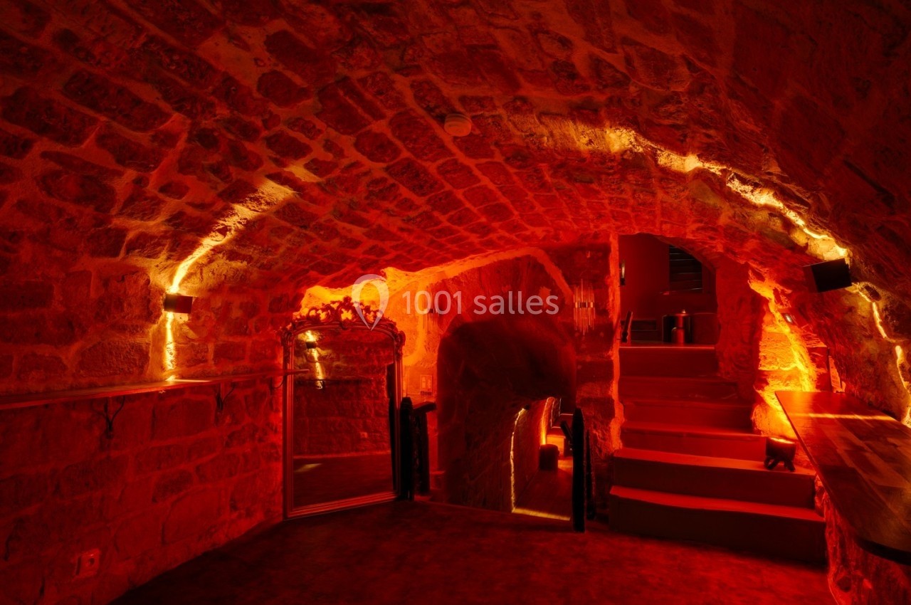 Cave voûtée en pierre éclairée par des lumières chaudes, avec un miroir et des escaliers en arrière-plan.