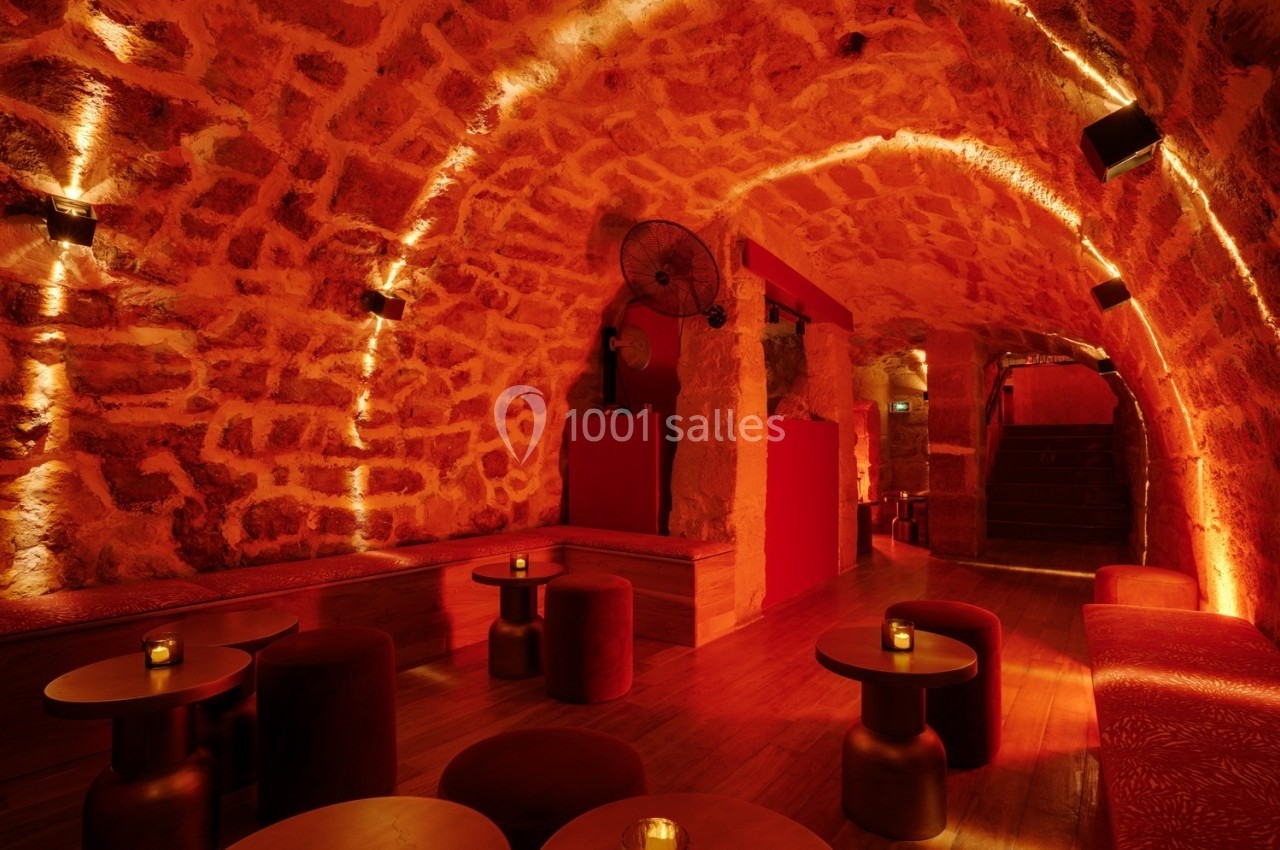 Salle voûtée en pierre éclairée par une lumière rouge tamisée, avec tables basses et sièges rembourrés.