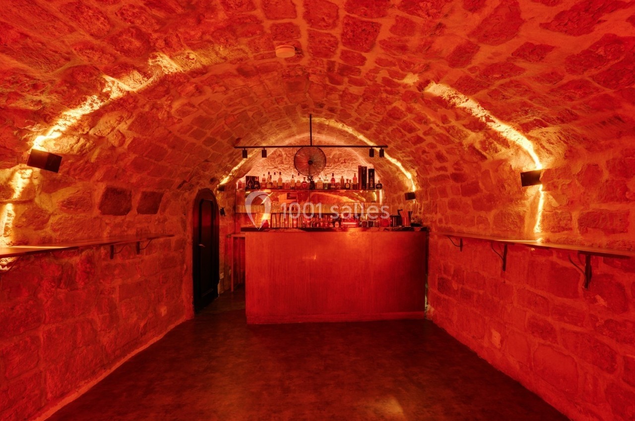 Bar en pierre voûtée avec éclairage rouge tamisé, comptoir en bois et étagères garnies de bouteilles.