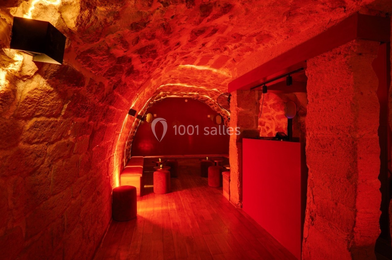 Salle voûtée en pierre éclairée de lumières rouges, avec des tabourets et une petite table en bois.