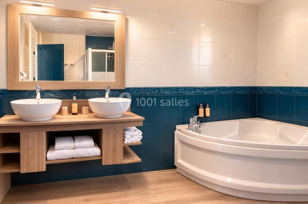 Salle de bain moderne avec double vasque, baignoire d'angle blanche, carrelage bleu et mobilier en bois clair.