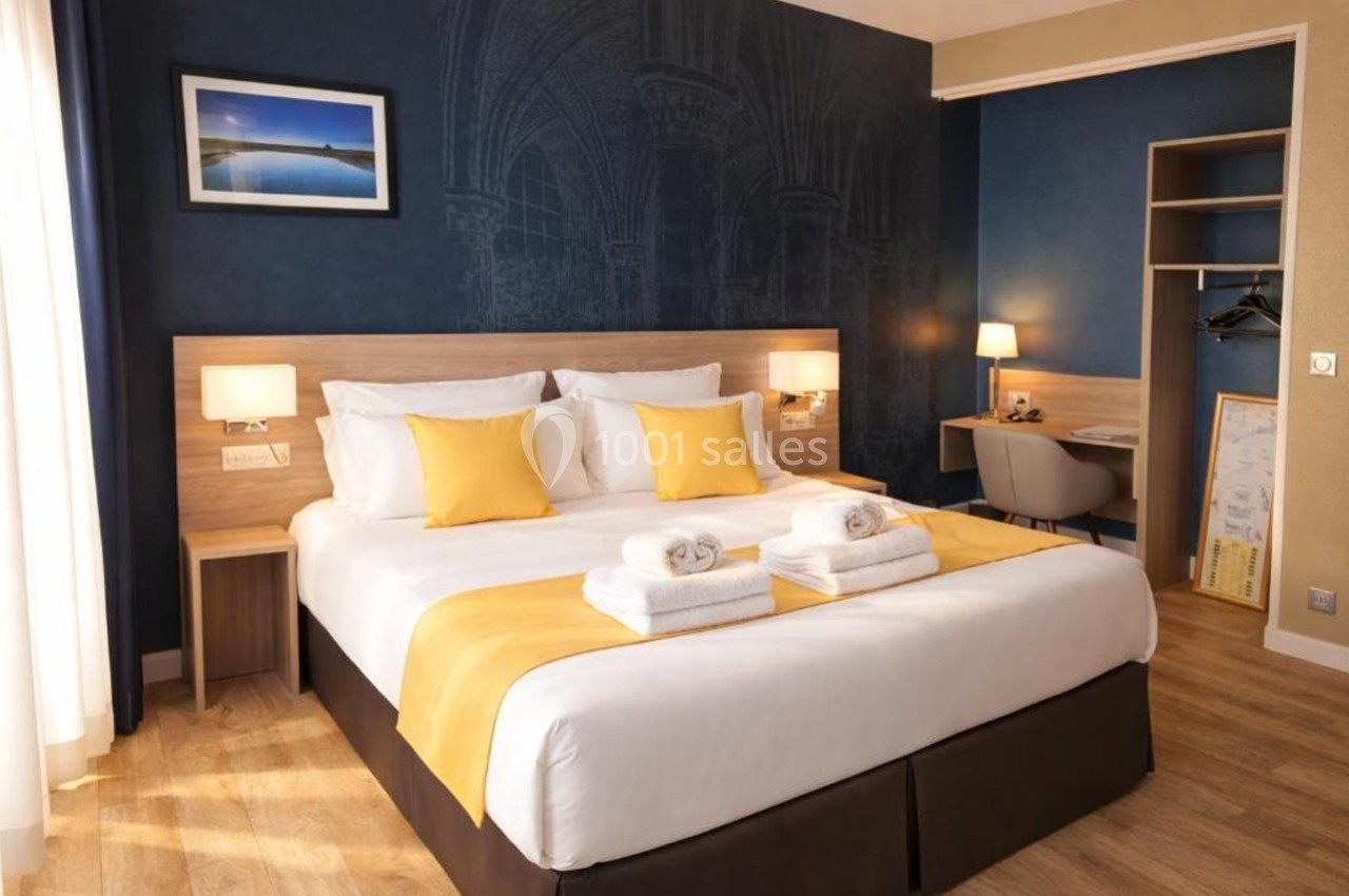 Chambre d'hôtel avec lit double, draps blancs, coussins jaunes, bureau, lampe allumée et décoration murale sombre.