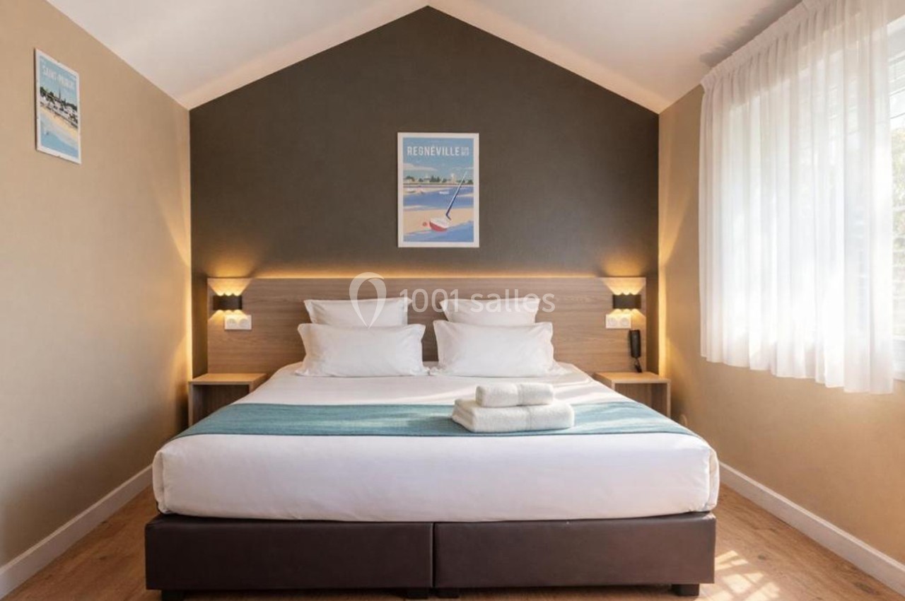 Chambre lumineuse avec un lit double, tête de lit en bois, deux lampes murales et une fenêtre avec rideaux blancs.