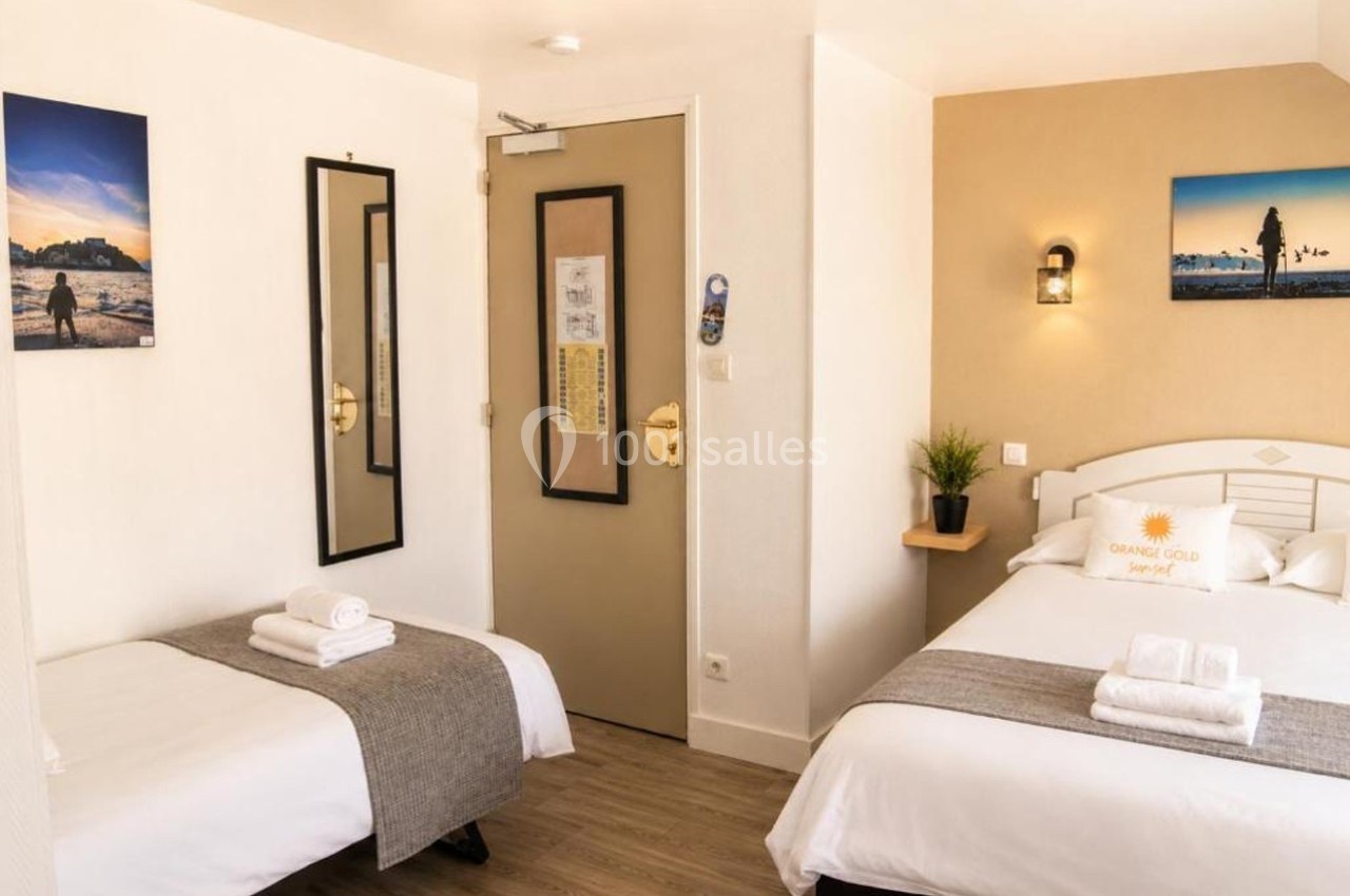 Chambre d'hôtel avec deux lits, décor sobre, murs clairs, tableaux, miroir, et porte beige avec des informations affichées.