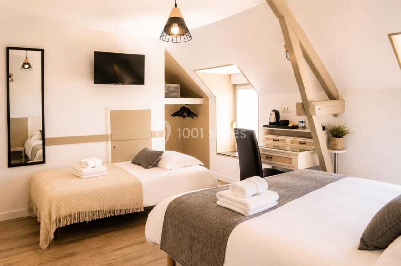 Chambre lumineuse avec deux lits, bureau, télévision murale, miroir et poutres apparentes.