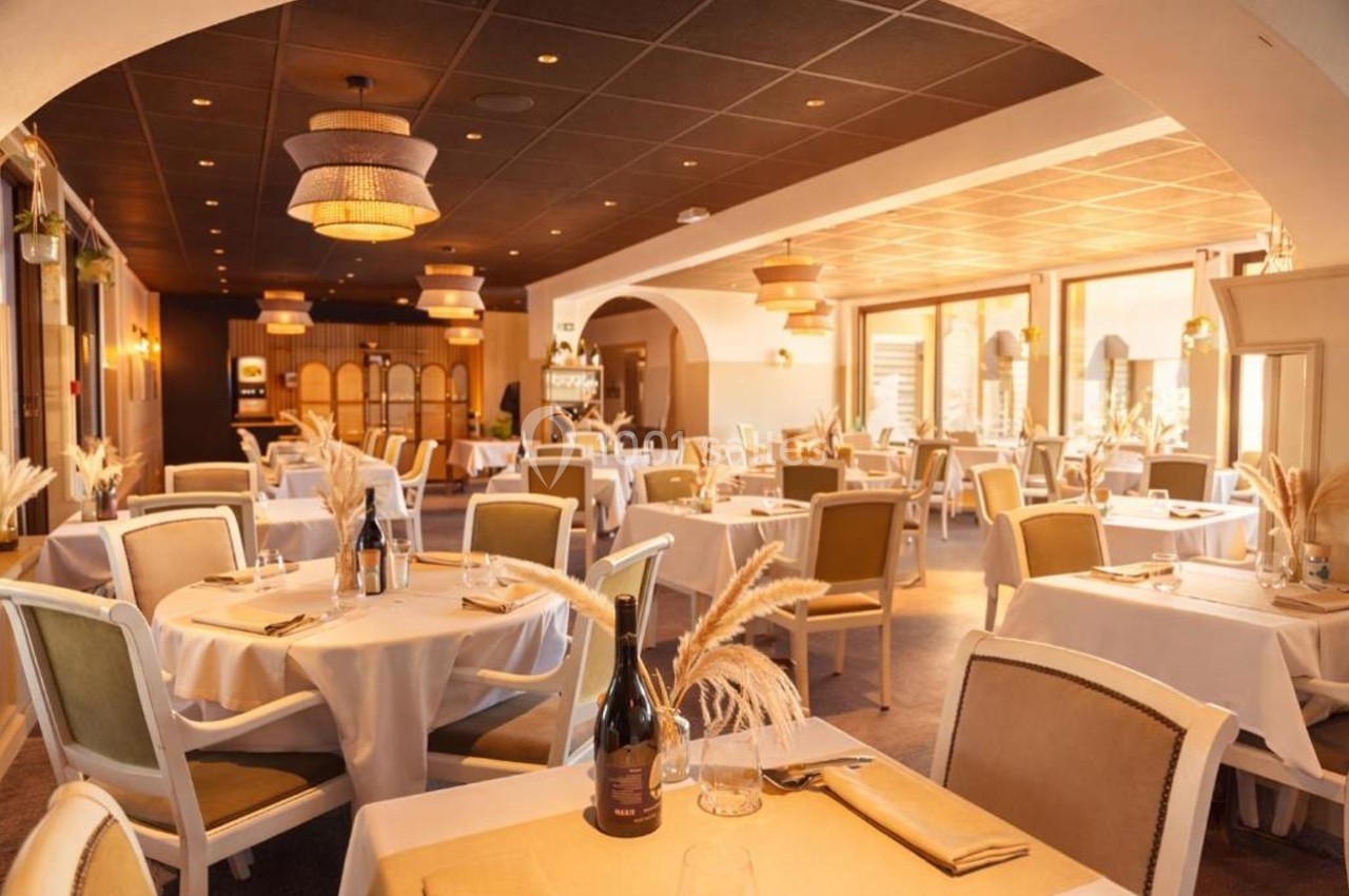 Salle de restaurant élégante avec tables dressées, chaises rembourrées et lumière naturelle entrant par de grandes fenêtres.