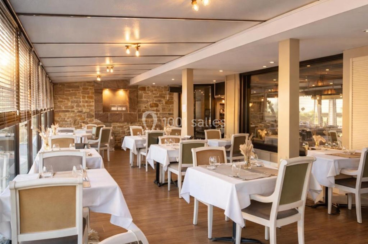 Salle de restaurant lumineuse avec tables dressées, chaises rembourrées et murs en pierre apparente.