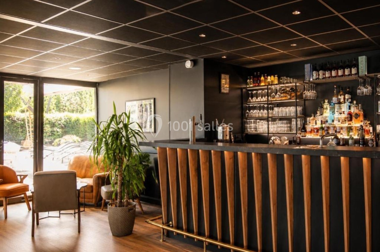 Bar moderne avec comptoir en bois, étagères de bouteilles et verres, fauteuils et plantes dans un espace lumineux.