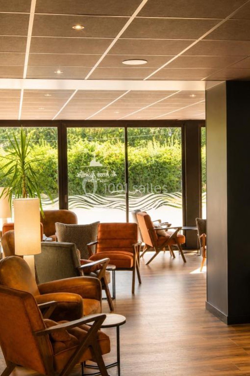 Espace lounge avec fauteuils en cuir, tables basses et grandes baies vitrées donnant sur un jardin verdoyant.