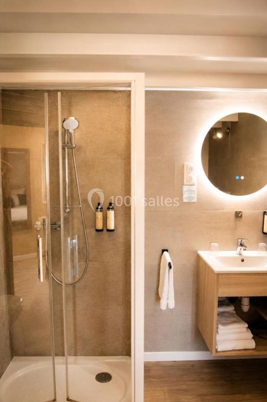 Salle de bain moderne avec douche vitrée, lavabo en bois, miroir rond éclairé et accessoires muraux.