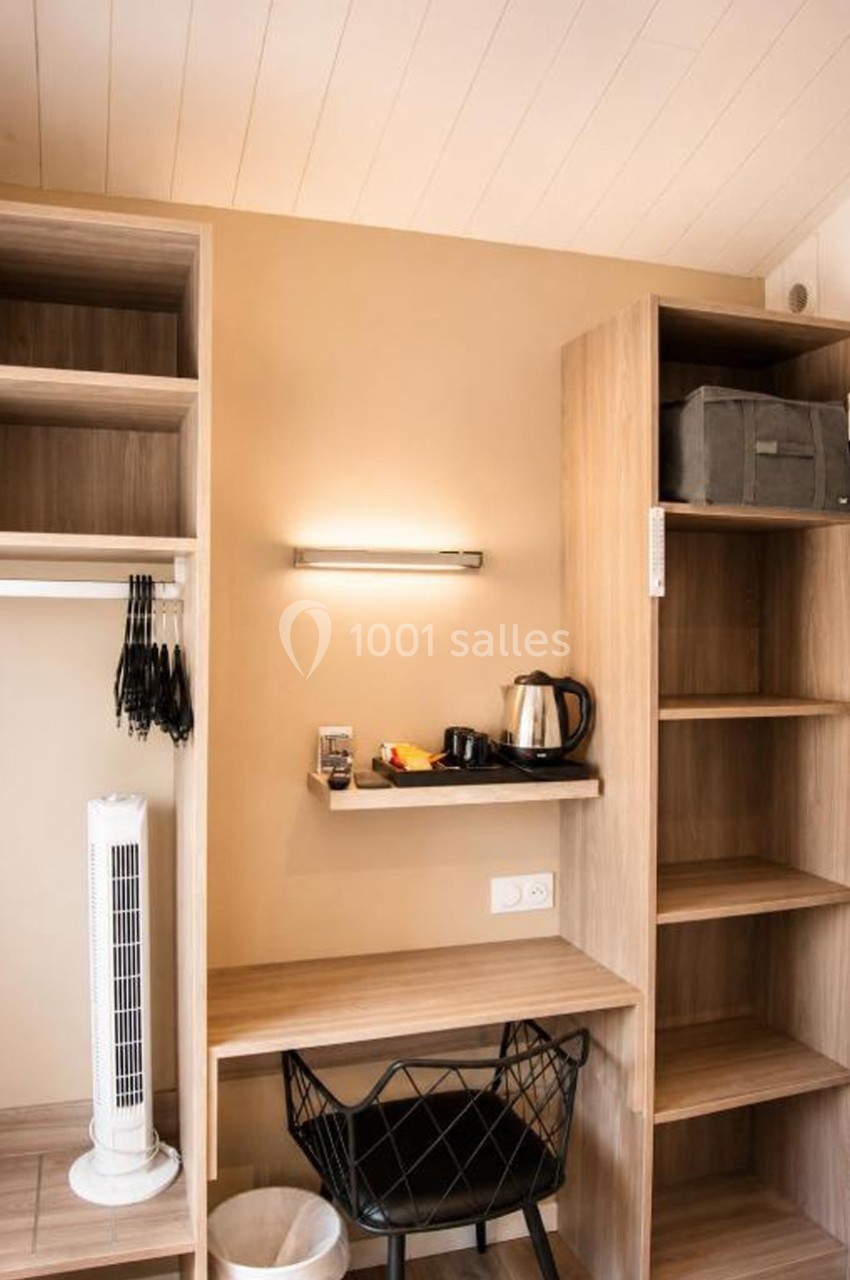 Bureau en bois avec chaise, étagères ouvertes, bouilloire, ventilateur sur pied et éclairage mural dans une pièce lumineuse.