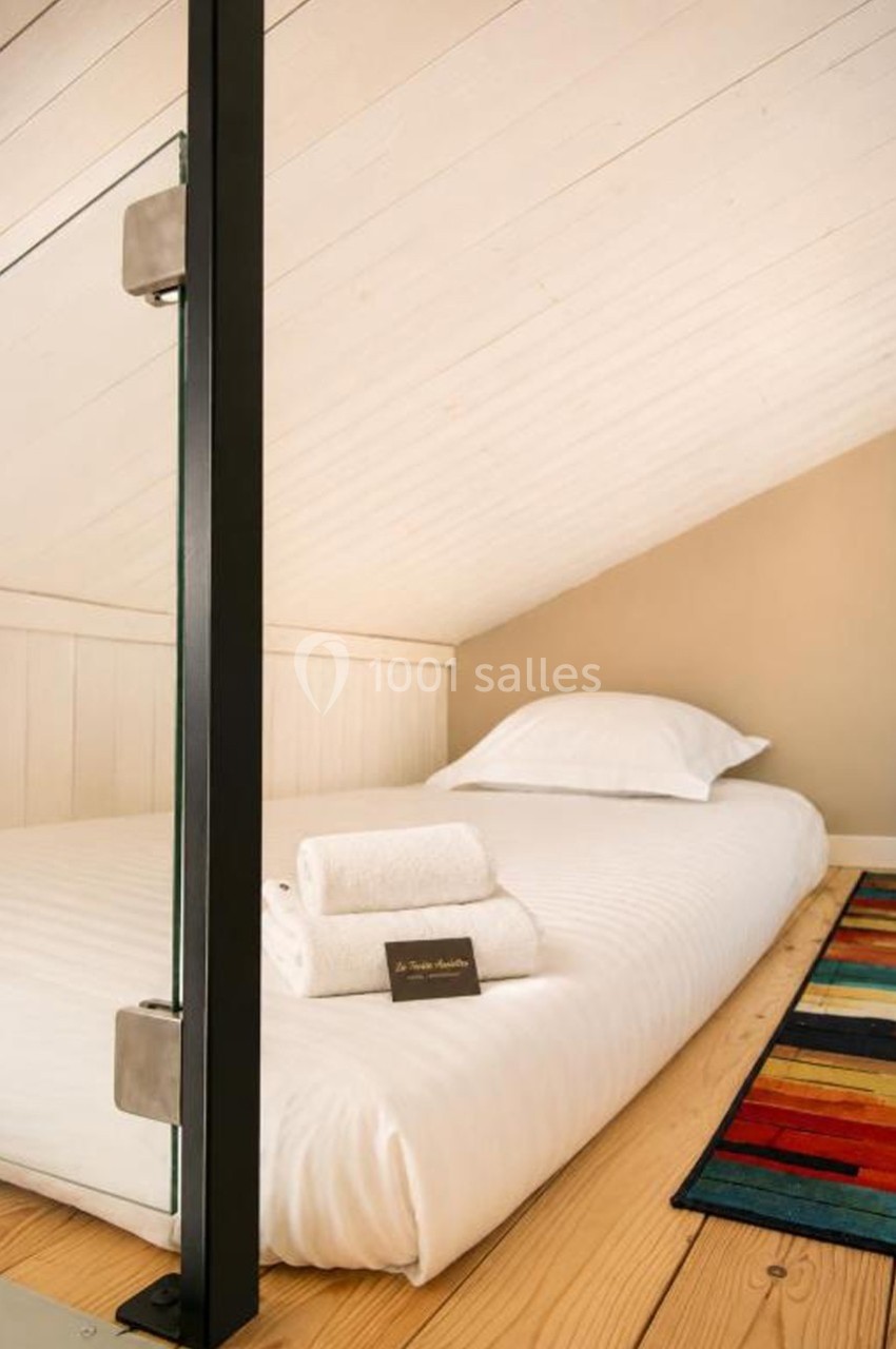 Lit simple sous un plafond mansardé, avec des serviettes pliées et une carte posées dessus, tapis coloré au sol.