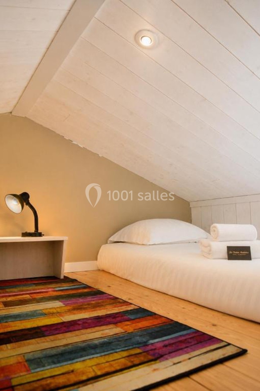 Lit simple avec draps blancs sous un plafond mansardé, lampe de chevet allumée et tapis coloré au sol.