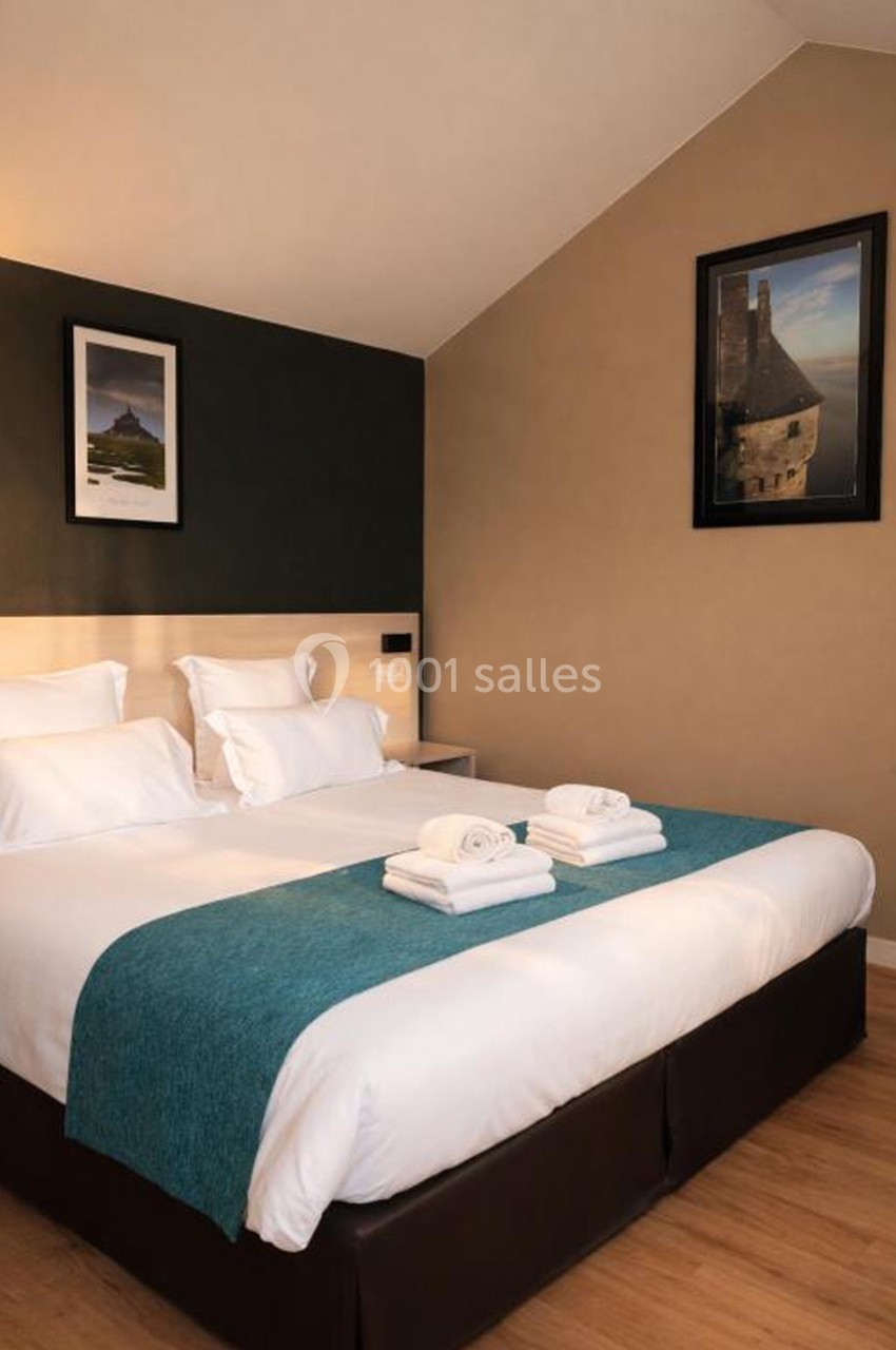 Lit double avec draps blancs, couverture turquoise et serviettes pliées, dans une chambre avec murs décorés de cadres.