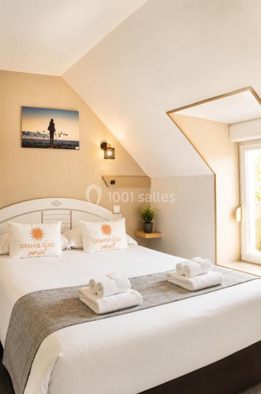 Lit double avec draps blancs, coussins décoratifs, serviettes pliées, dans une chambre lumineuse avec fenêtre et décoration…