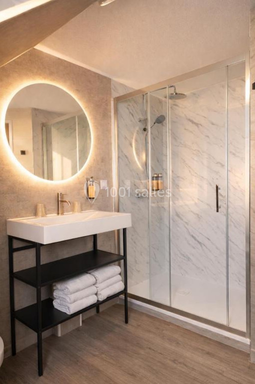 Salle de bain moderne avec douche à paroi vitrée, lavabo sur meuble noir et miroir rond éclairé.