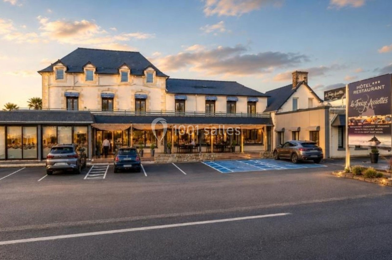 Façade d'un hôtel-restaurant avec parking, terrasse et enseigne visible sous un ciel partiellement nuageux.