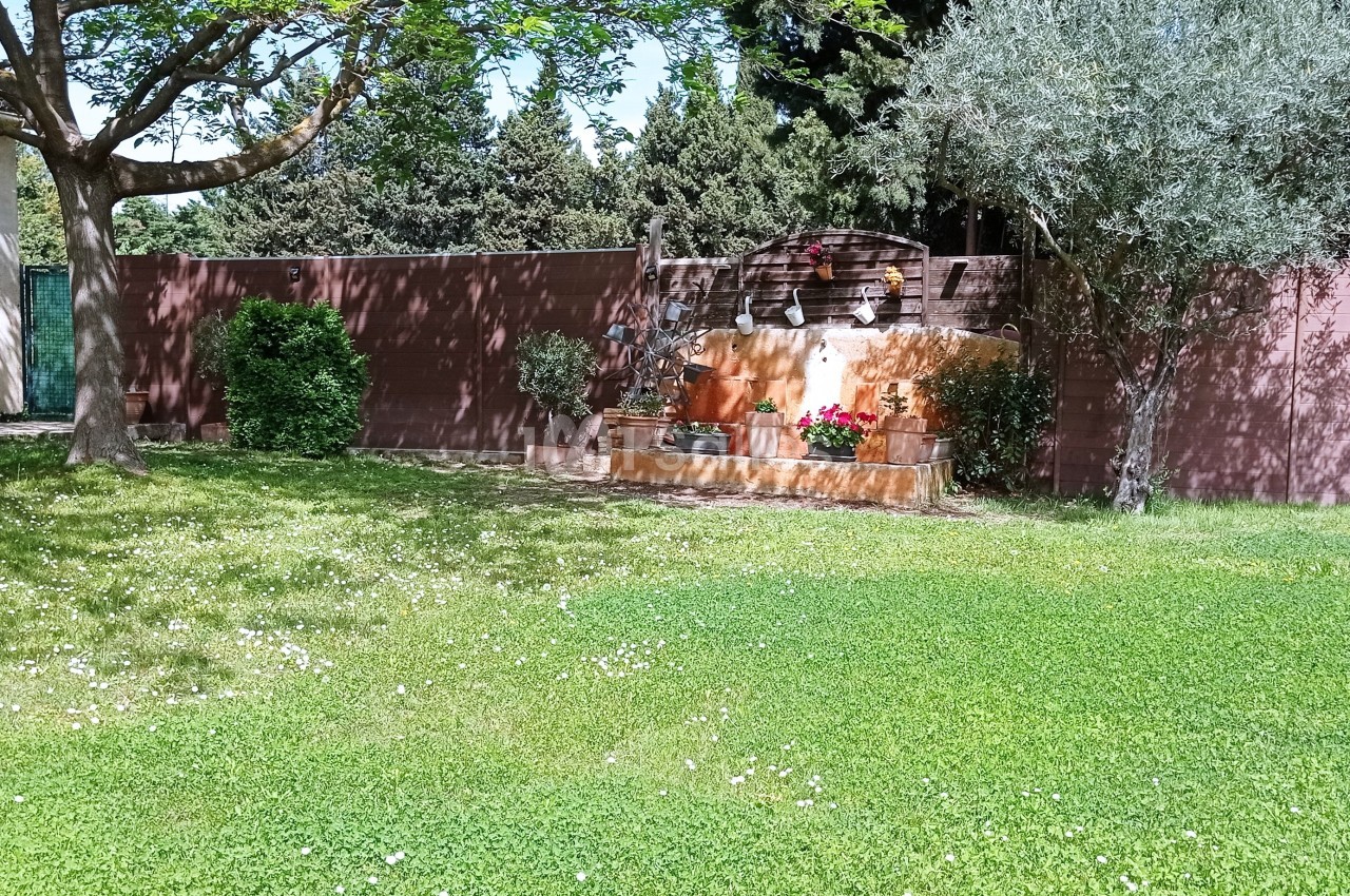 Jardin avec pelouse, fontaine en pierre ornée de pots de fleurs, arbres et mur en arrière-plan.