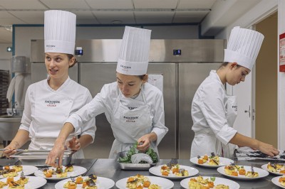 Trois cheffes en tenue blanche dressent des assiettes gastronomiques dans une cuisine professionnelle.