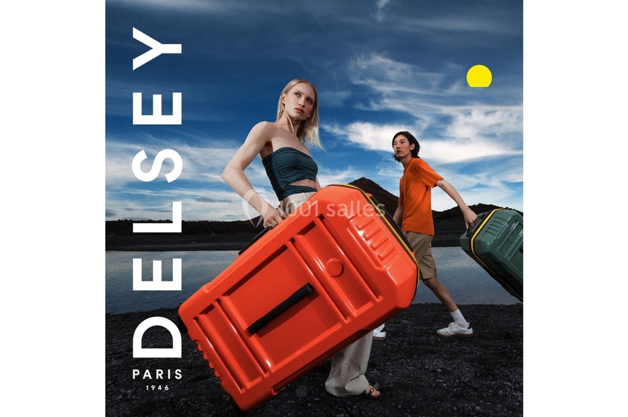 Deux personnes transportent des valises colorées dans un paysage extérieur sous un ciel partiellement nuageux.