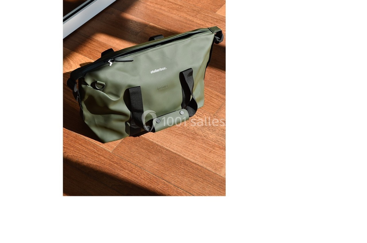 Sac vert olive posé sur un sol en bois, avec des poignées noires et une fermeture éclair visible.