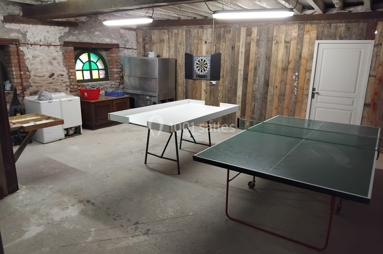 Salle de loisirs avec table de ping-pong, table de jeu blanche, cible de fléchettes et murs en bois et pierre.
