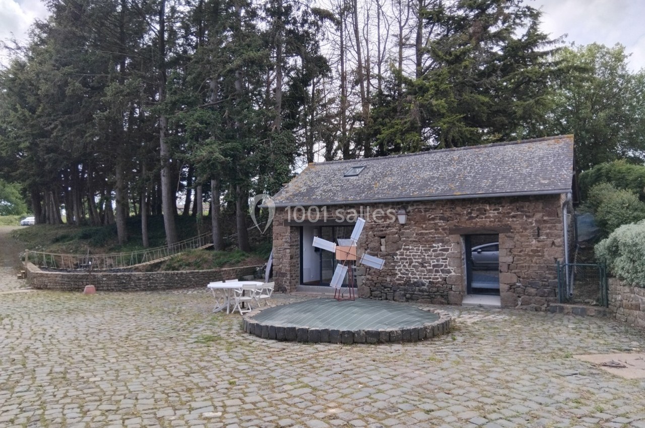 Bâtiment en pierre avec toit en ardoise, petite terrasse en bois, tables blanches et arbres en arrière-plan.