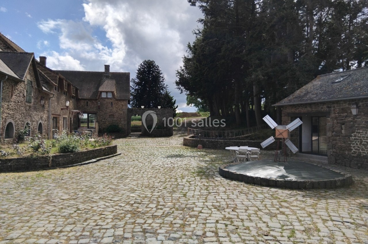 Cour pavée avec bâtiments en pierre, un petit moulin décoratif et des arbres en arrière-plan sous un ciel partiellement…