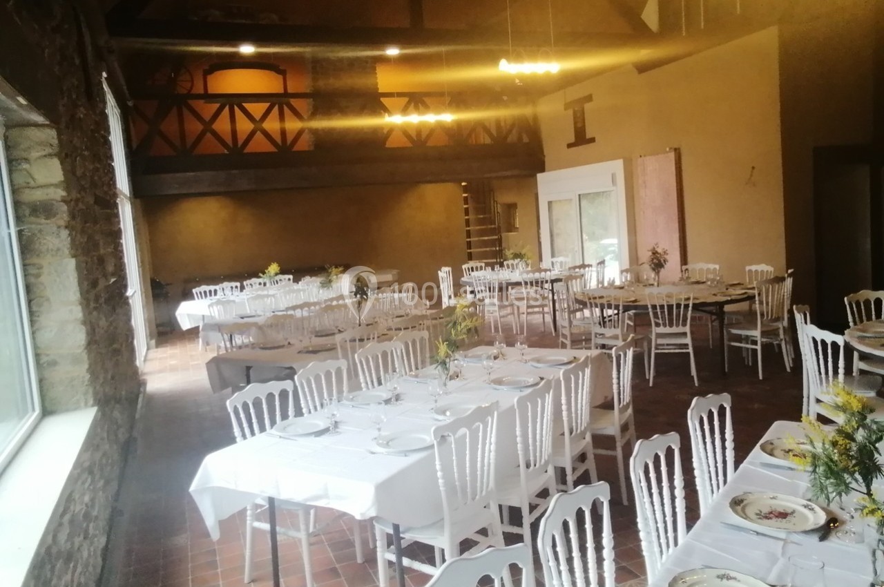 Salle de réception avec tables dressées, nappes blanches, chaises en bois et décoration florale, éclairée par la lumière…