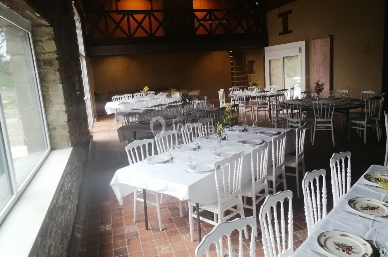 Salle de réception avec tables dressées, nappes blanches et chaises, éclairée par une grande baie vitrée.