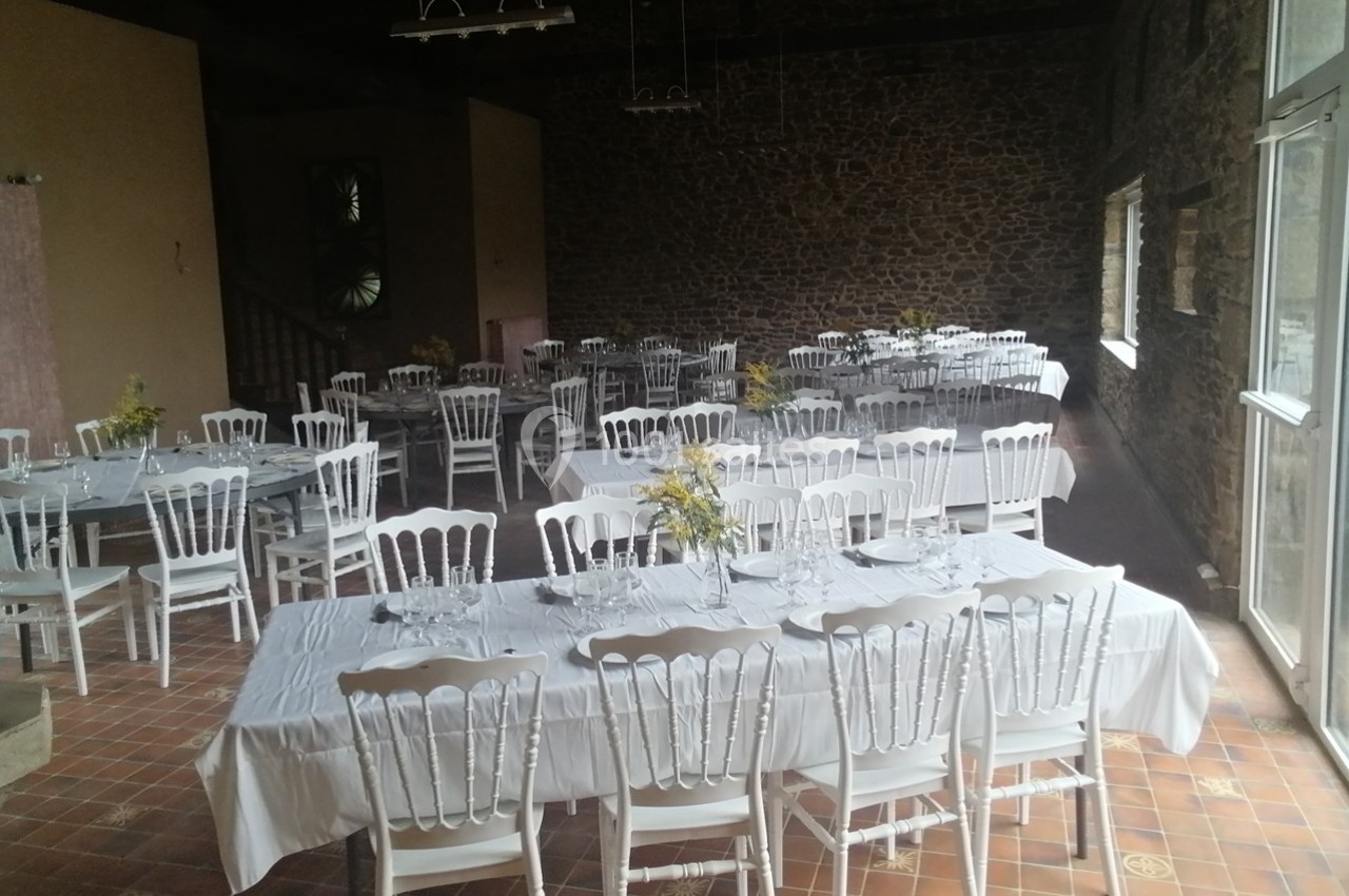 Salle de réception avec des tables dressées, nappes blanches, chaises blanches et décoration florale sur un sol en carreaux.