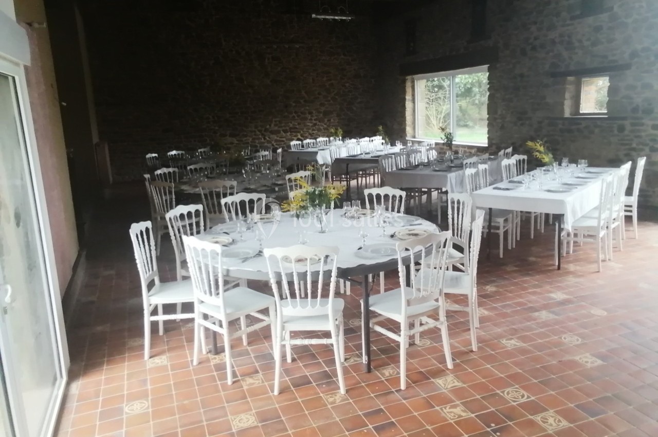 Salle de réception avec tables rondes et rectangulaires dressées, chaises blanches et décoration florale.