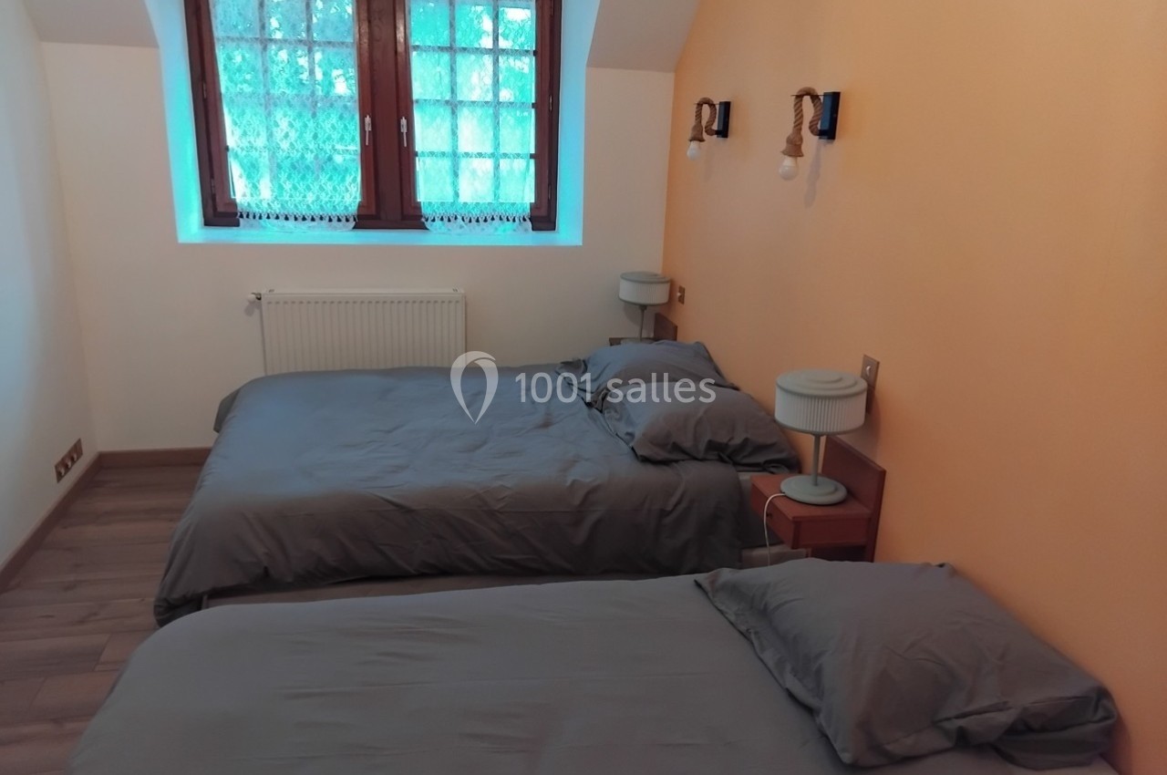 Deux lits simples avec draps gris dans une chambre aux murs orange, éclairée par une fenêtre avec vue sur des arbres.