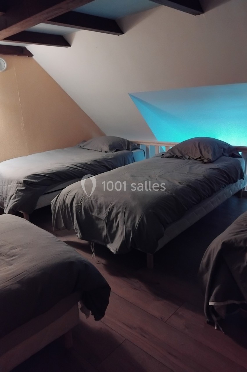 Quatre lits simples alignés dans une chambre mansardée avec éclairage tamisé et poutres apparentes.