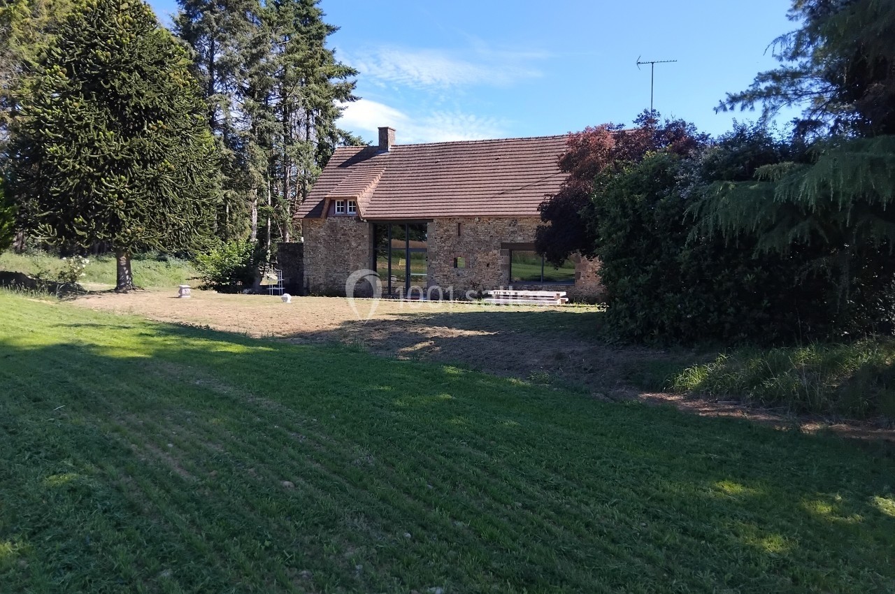 Maison en pierre avec toit en tuiles entourée de pelouse, d'arbres et d'un ciel dégagé.