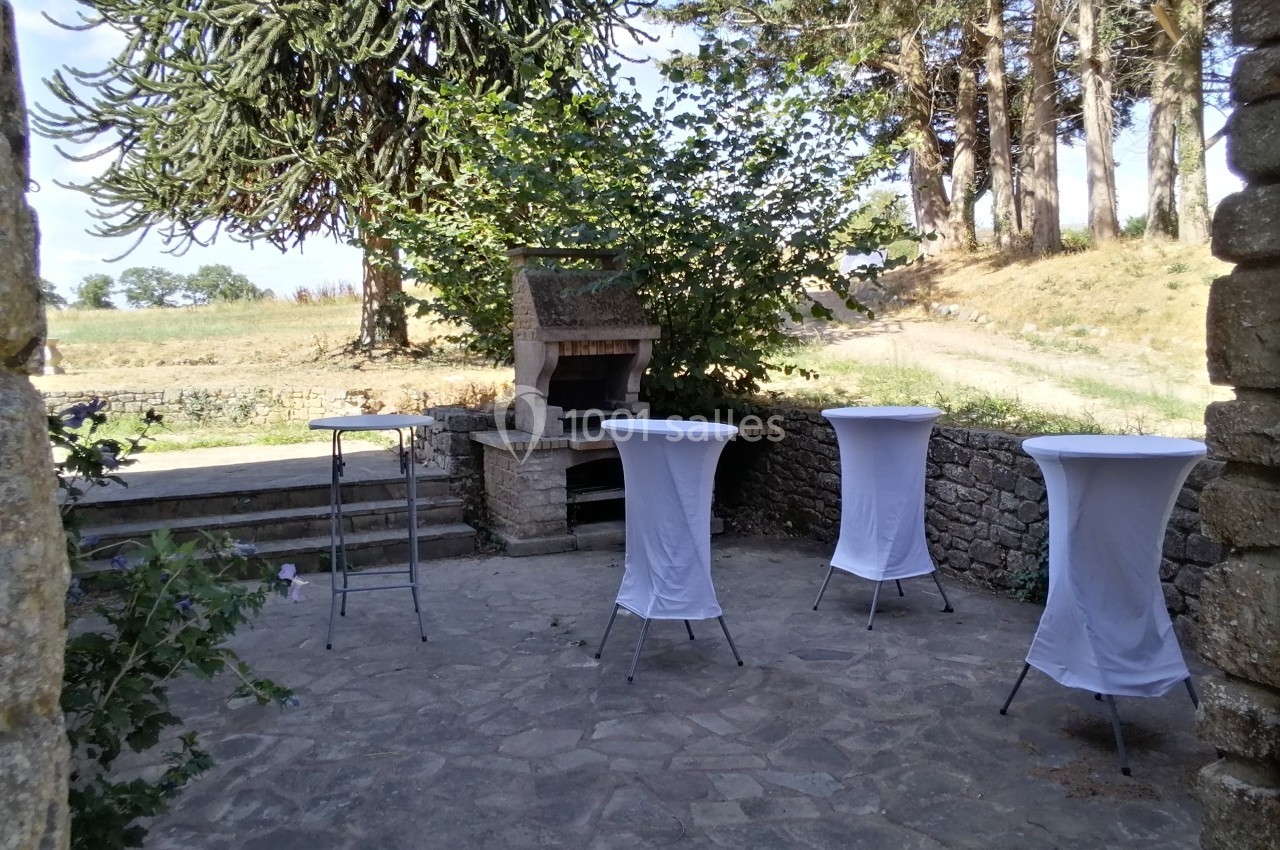 Terrasse en pierre avec un barbecue en pierre, des tables hautes couvertes de nappes blanches et des arbres en arrière-plan.