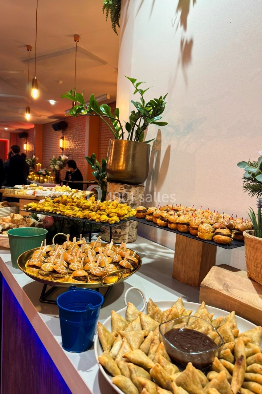 Buffet avec assortiment de bouchées salées, samoussas, mini-burgers et sauces, dans une salle décorée de plantes.