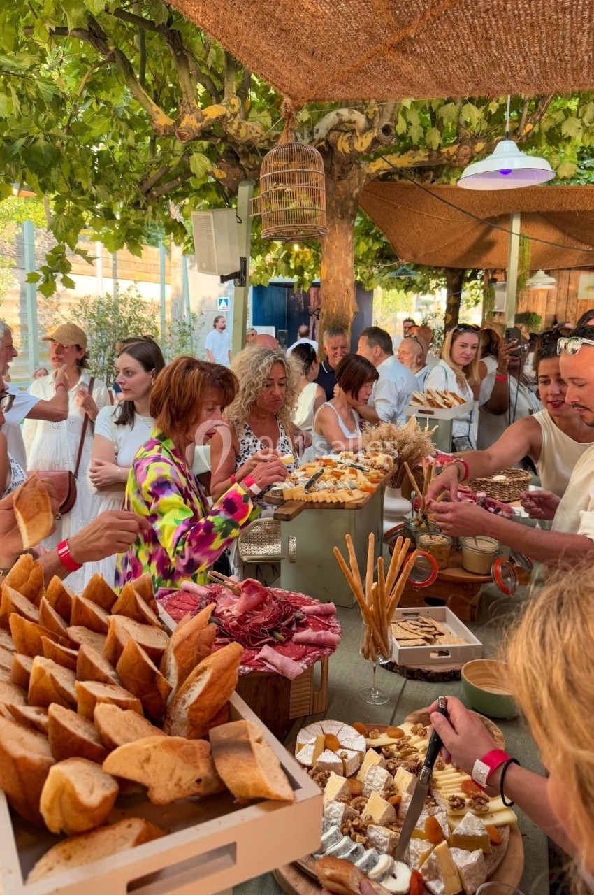 Groupe de personnes partageant un buffet en plein air avec charcuterie, fromages et pâtisseries sous un auvent ombragé.