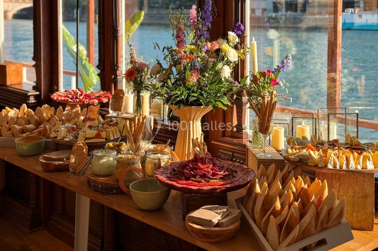 Table garnie de charcuteries, pains, fromages et fleurs, disposée près de grandes fenêtres donnant sur une rivière.