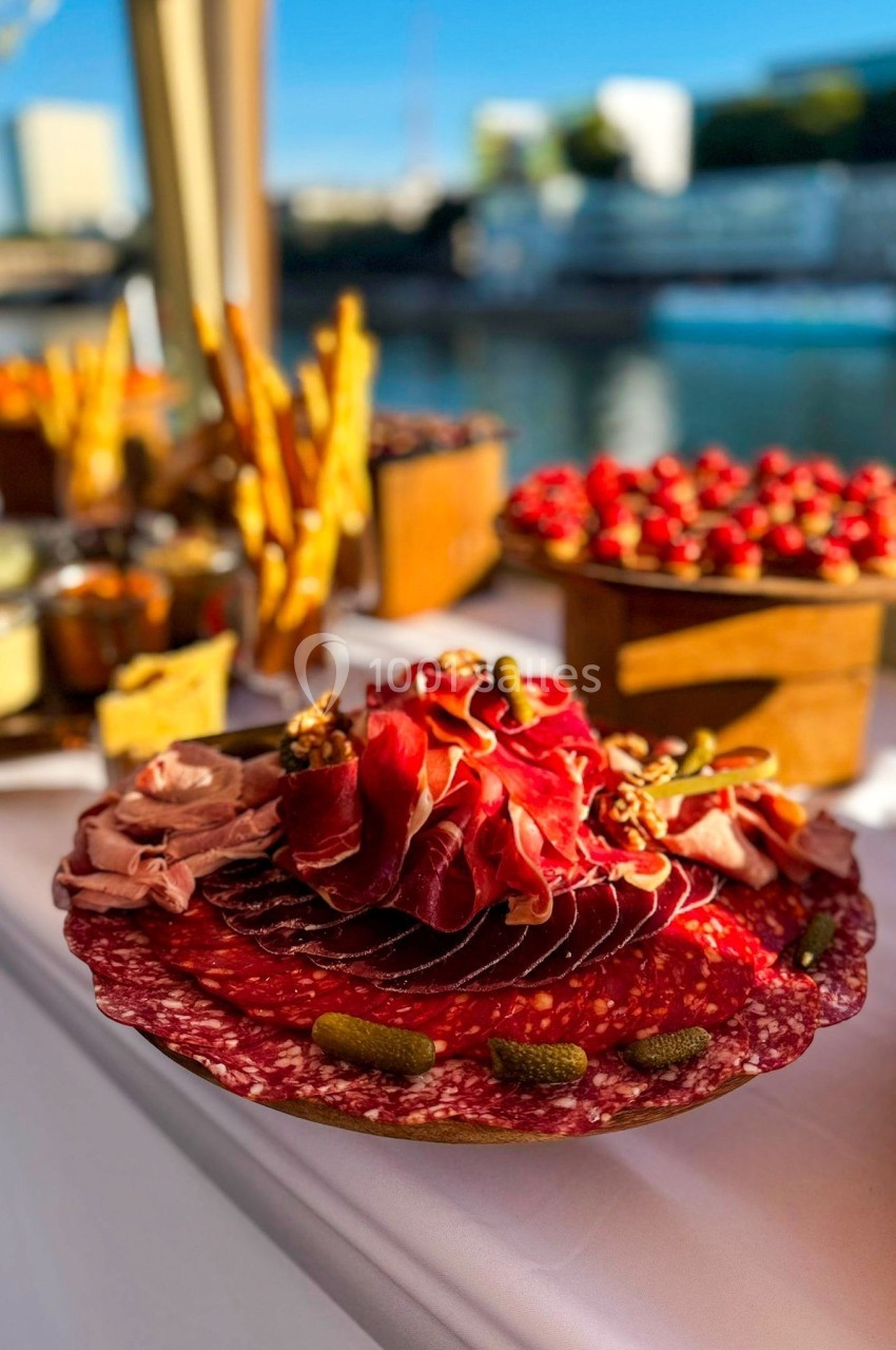 Plateau de charcuterie avec tranches de jambon, saucisson, cornichons et noix, disposé sur une table en extérieur.