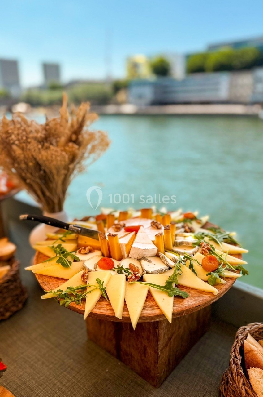 Plateau de fromages variés décoré de fruits secs et roquette, présenté sur une table en bord de rivière.