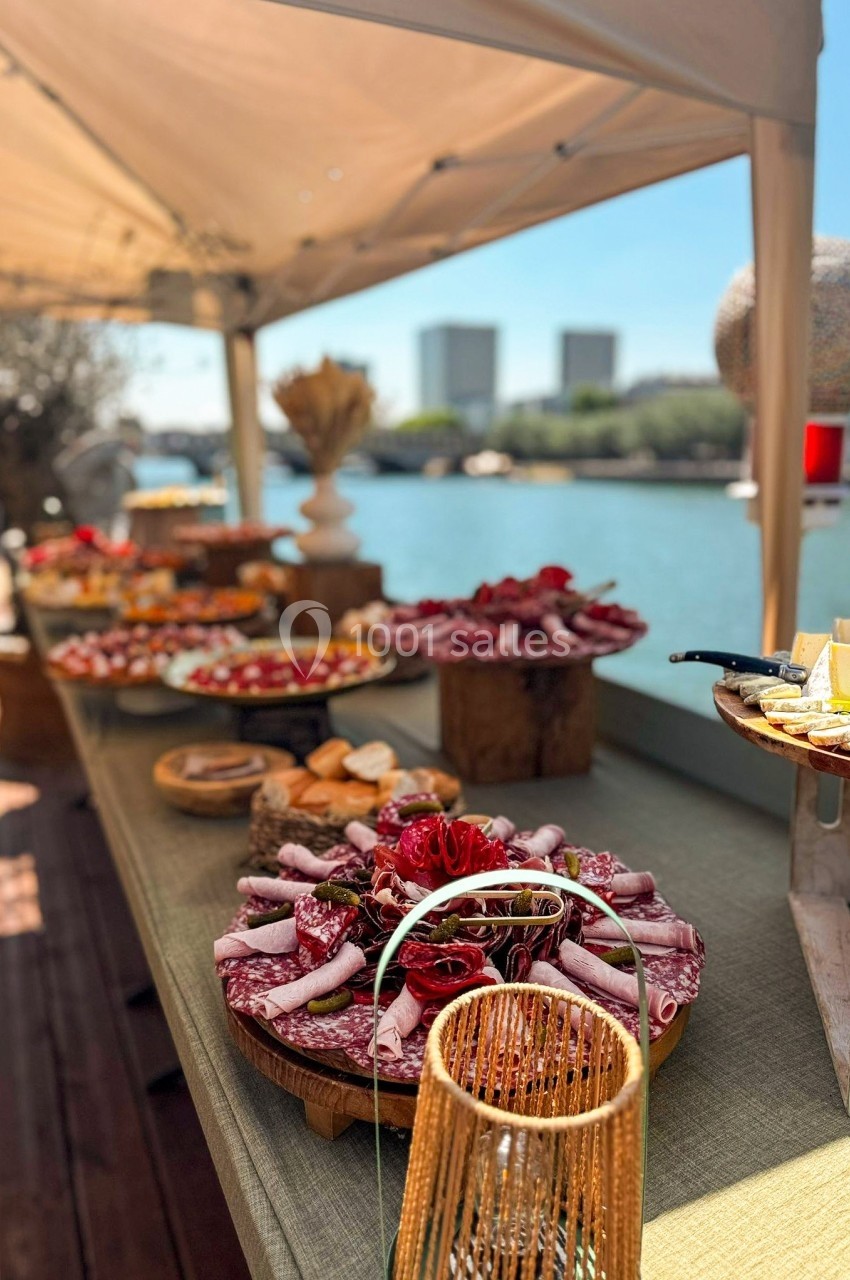 Table garnie de plats variés, incluant charcuterie et fromages, installée en extérieur près d'un plan d'eau.