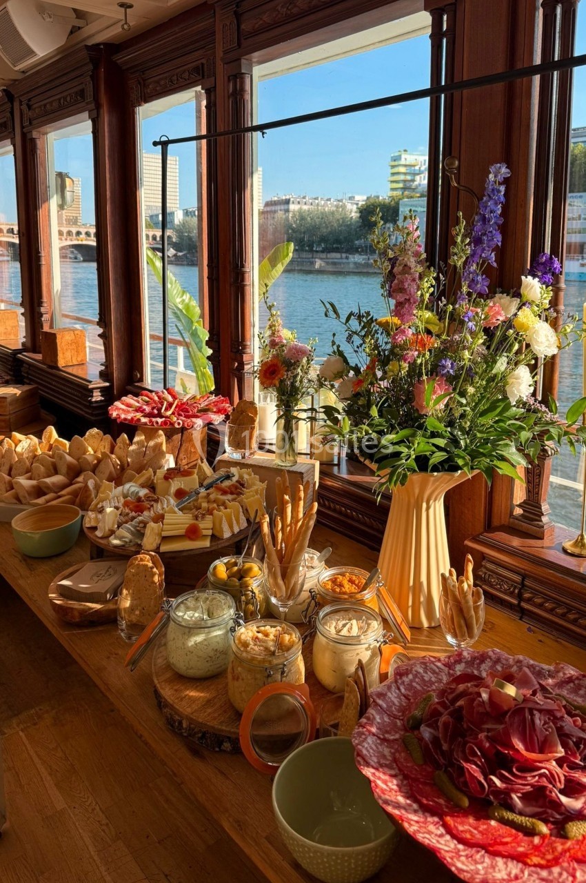 Buffet avec charcuterie, pains, dips et fleurs, installé dans une salle lumineuse avec vue sur une rivière.