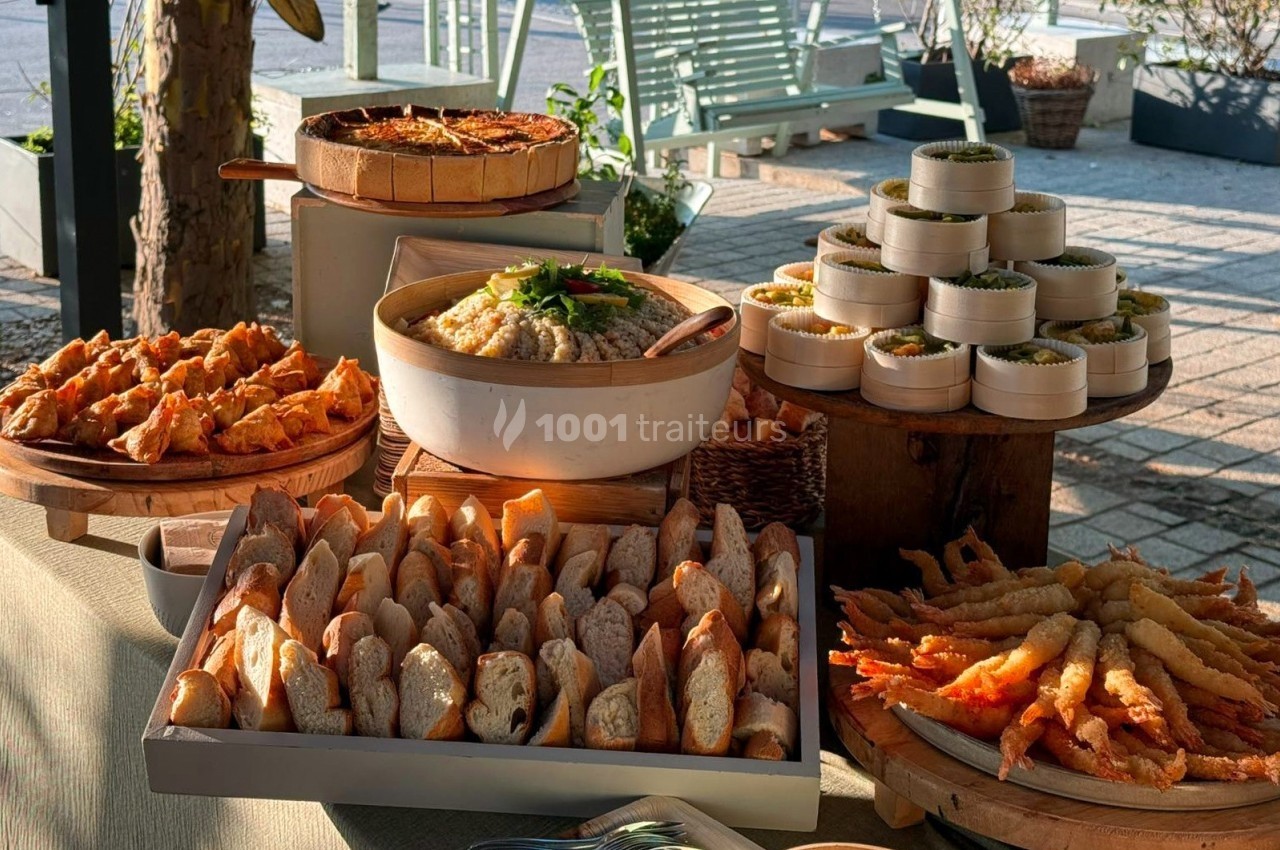 Buffet en extérieur avec pain, quiches, beignets de crevettes, mezzés et autres plats disposés sur une table en bois.