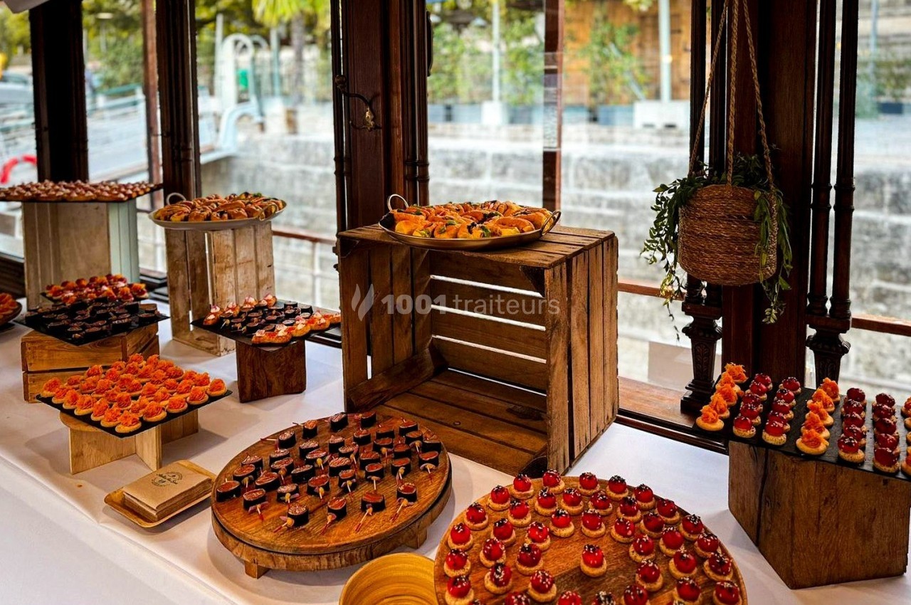 Buffet de petites bouchées colorées disposées sur des présentoirs en bois près de grandes fenêtres donnant sur l'extérieur.
