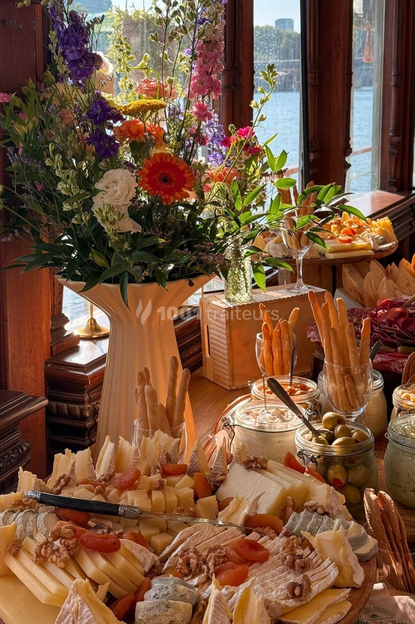 Plateau de fromages variés avec fruits secs, olives et pain, disposé près d'un bouquet de fleurs et d'une fenêtre…