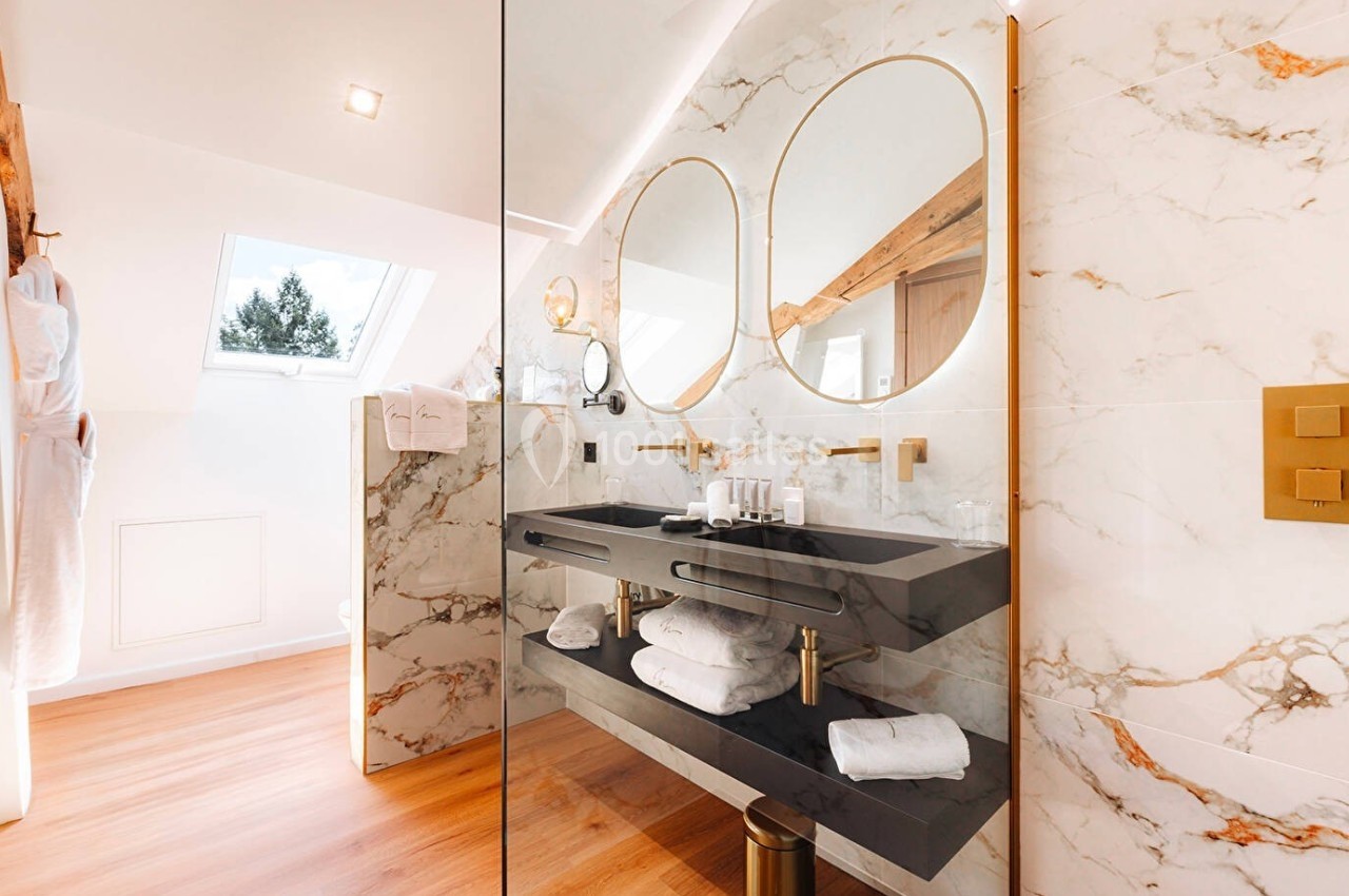 Salle de bain moderne avec double vasque noire, miroirs ronds, finitions dorées et murs en marbre clair.